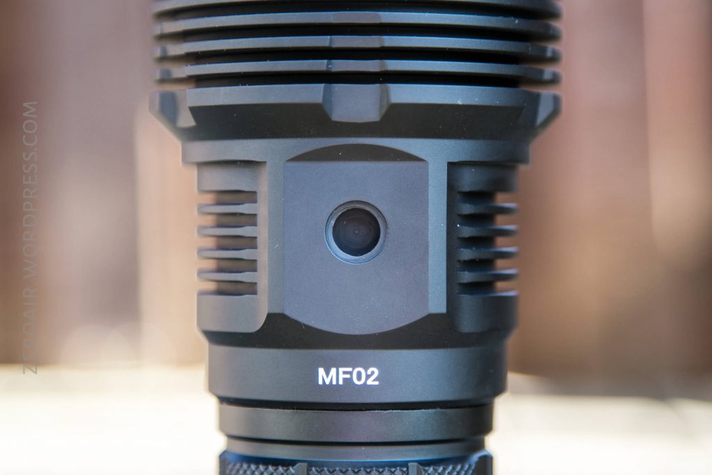 Close-up view of a black cylindrical device labeled MF02 with ridged edges and a central circular feature, possibly a lens or sensor, set against a blurred background.