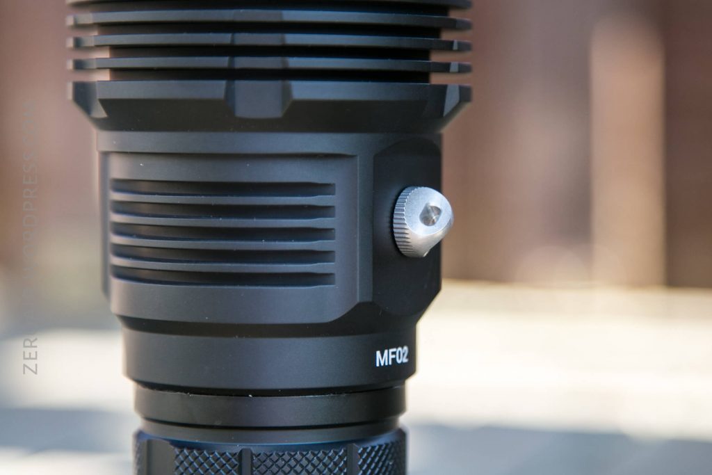 Close-up of a black cylindrical device with ribbed texture and a metallic screw on the side, labeled MF02. The background is blurred.