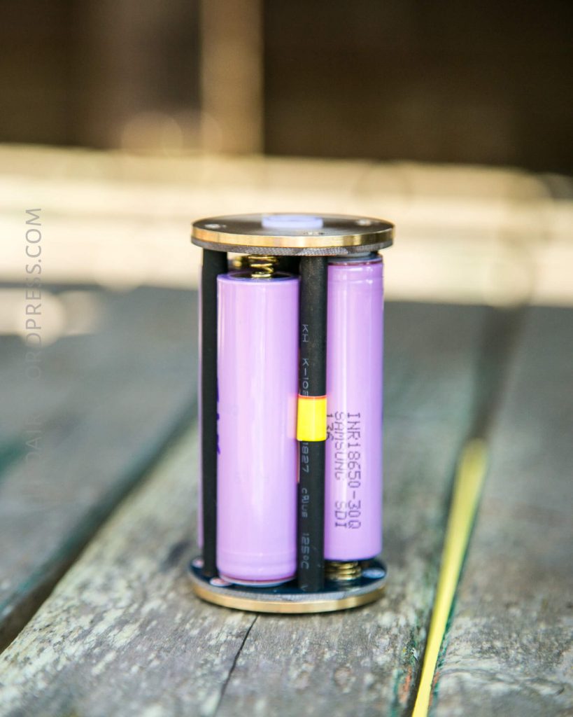 A cylindrical battery holder with two purple batteries inserted vertically, connected by black rods and metal end caps, sits on a wooden surface.