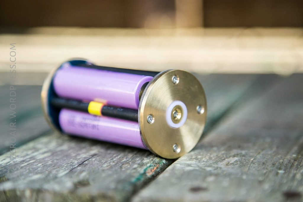 A cylindrical battery holder with two purple rechargeable batteries inside, metal end caps, and black connecting rods sits on a weathered wooden surface.