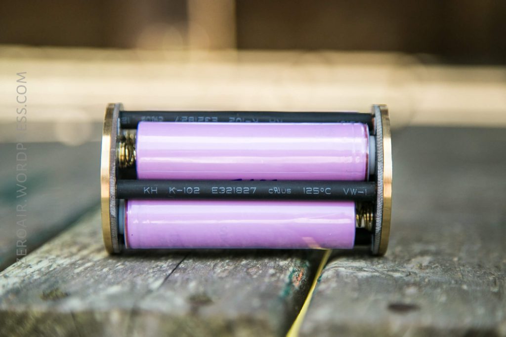 Two cylindrical pink batteries are placed horizontally in a black and metal battery holder, which is lying on its side on a wooden surface. Text and numbers are visible on the batteries and the holder.