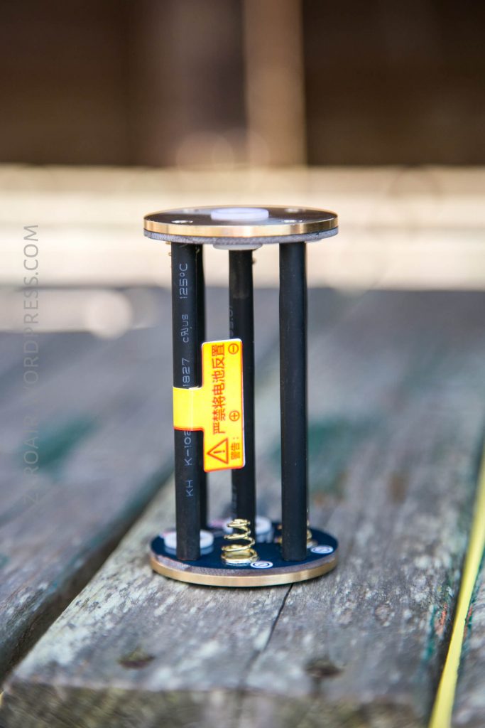 A close-up of a cylindrical electronic component with two circular metal plates connected by four black rods, positioned upright on a wooden surface. A yellow caution label is attached to one of the rods.