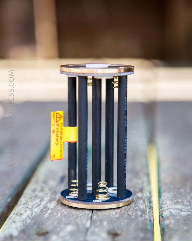 A cylindrical device with black vertical rods, metal top and bottom plates, visible springs at the base, and a yellow caution label attached to one rod, placed on a wooden surface.