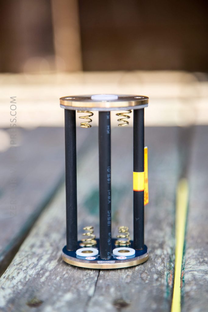 A cylindrical object with two round metal plates connected by four black rods, each with a metal spring inside. The object is standing vertically on a wooden surface.