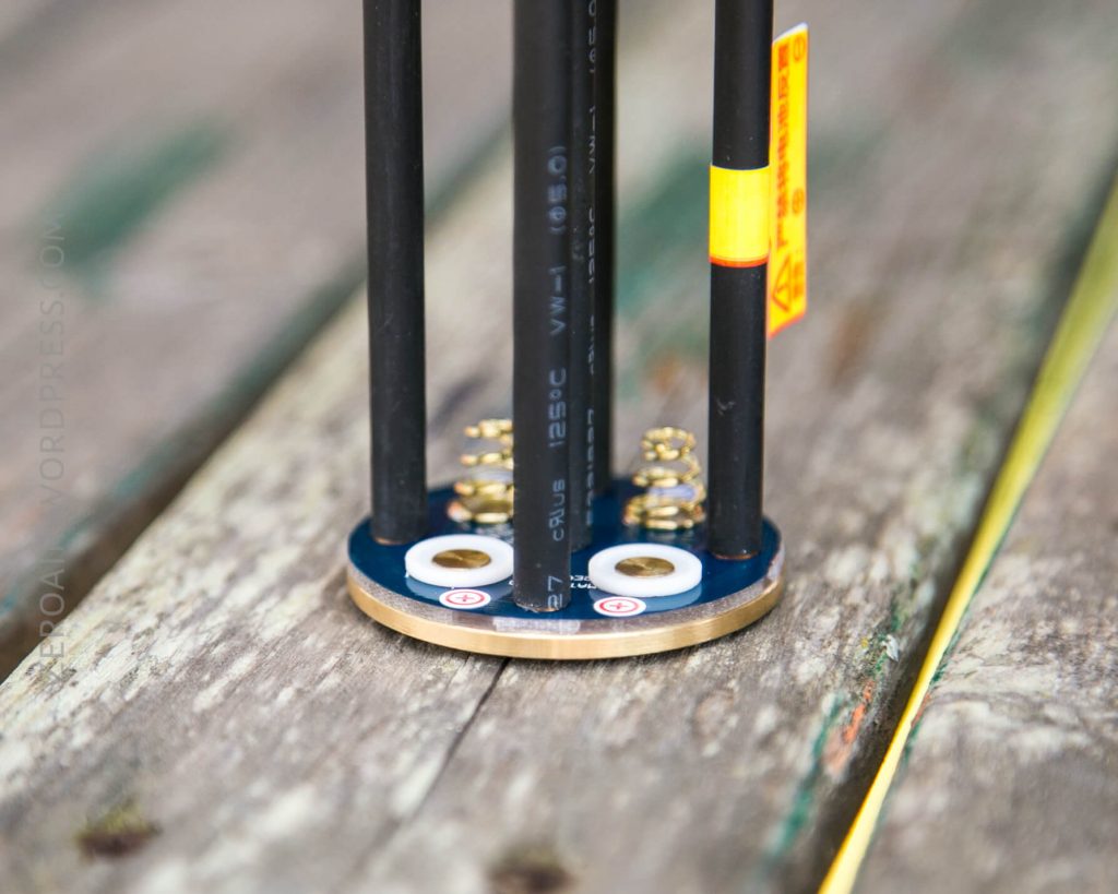 Close-up of the base of a mechanical or electronic device with four vertical rods attached to a circular metal plate, two springs, and wiring on a weathered wooden surface.