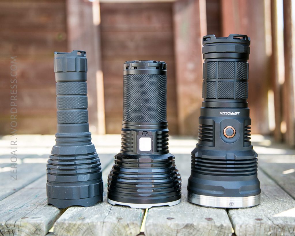 Three large black flashlights stand upright in a row on a wooden surface. The background features wooden paneling with sunlight casting shadows. The flashlights have different designs and button placements.