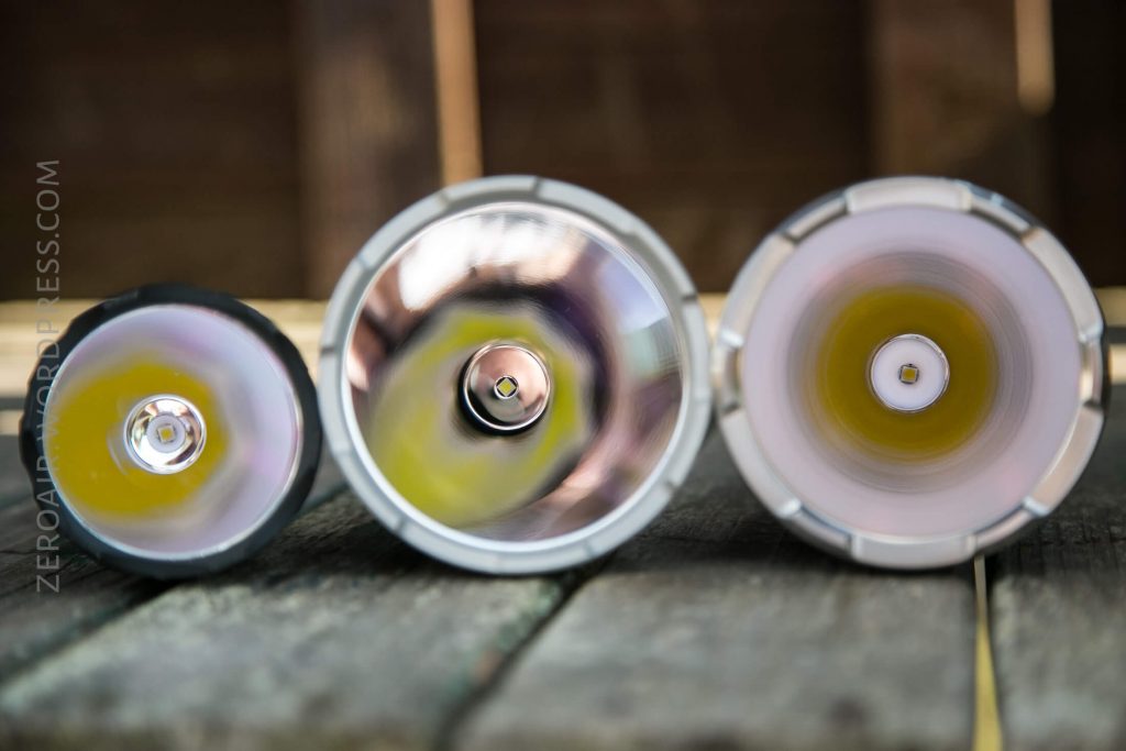Three flashlight heads are shown side by side on a wooden surface, photographed from above to display the reflectors and LED emitters inside each flashlight. The background is blurred.