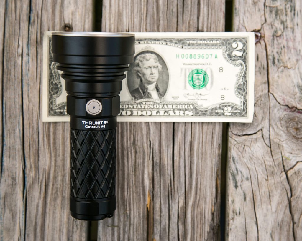 A black ThruNite Catapult V6 flashlight is placed vertically on top of a two-dollar bill, which lies flat on a wooden surface. The flashlight is shorter than the length of the bill.