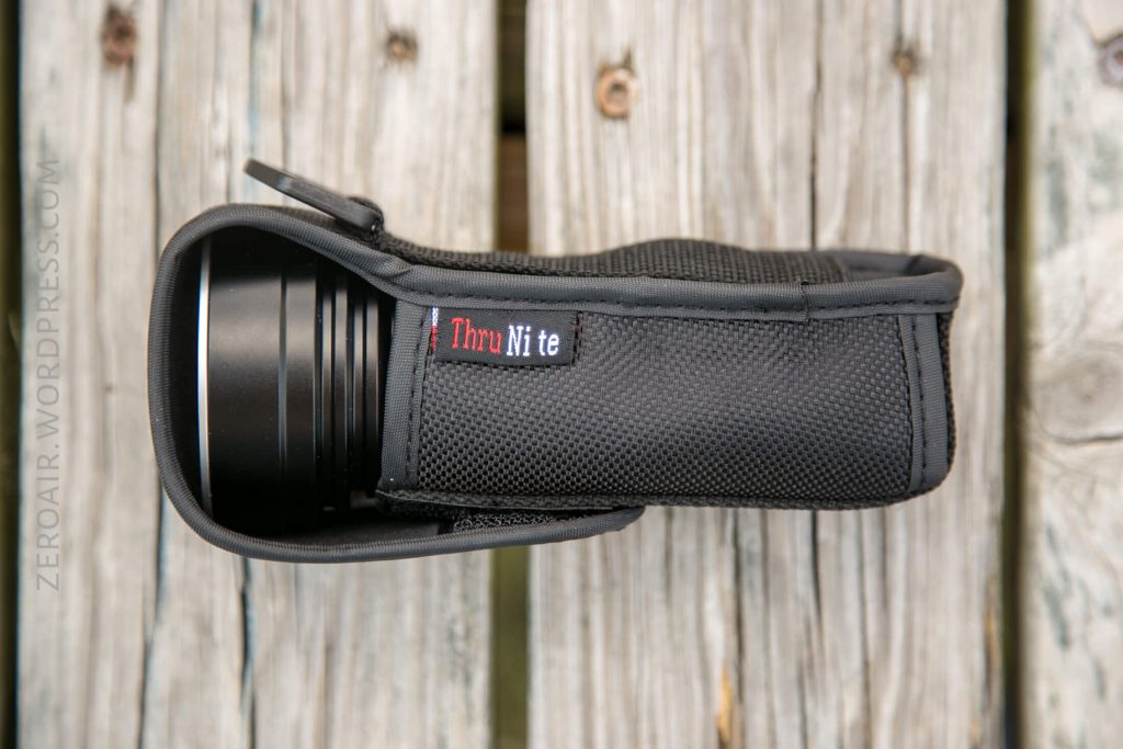 A black ThruNite flashlight in a textured fabric case is placed on a weathered wooden surface. The ThruNite brand label is visible on the side of the case.