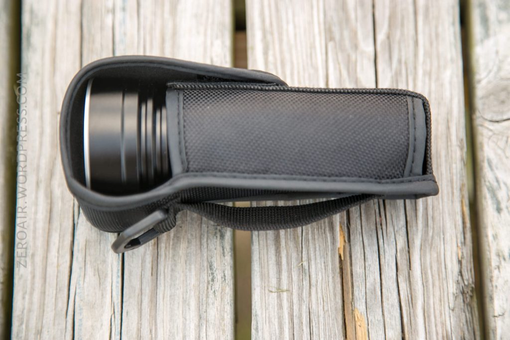 A black flashlight in a fitted black fabric holster with a strap, resting on a weathered wooden surface. ZEROAIR.WORDPRESS.COM is visible along the left side of the image.