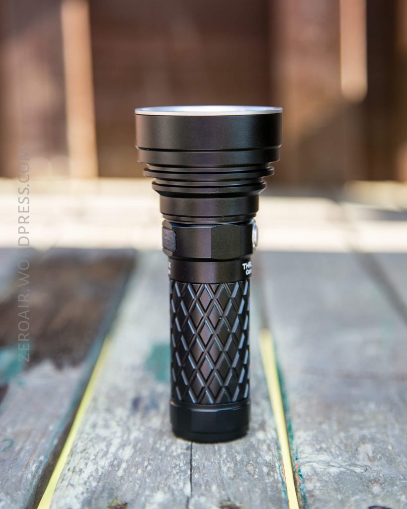 A black flashlight with a textured grip stands upright on a weathered wooden surface, with a blurred brown background.