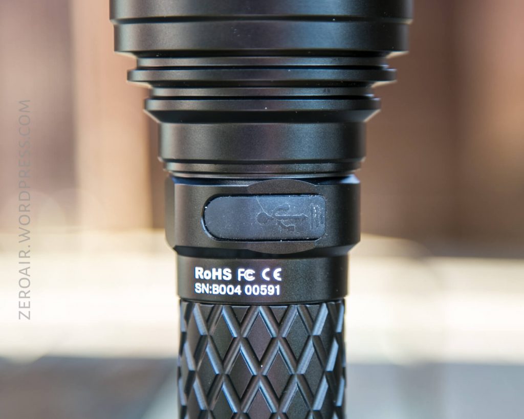 Close-up of a black flashlight showing a covered USB charging port, a textured grip, and white printed certification markings including RoHS, FCC, and a serial number. The background is out of focus.