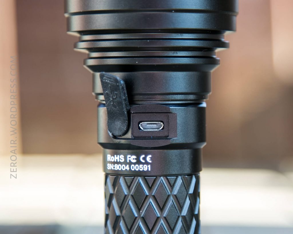 A close-up of a black flashlight shows a micro-USB charging port with a rubber cover partially open. The flashlight has a textured grip and visible certification markings including RoHS, FC, CE, and a serial number.