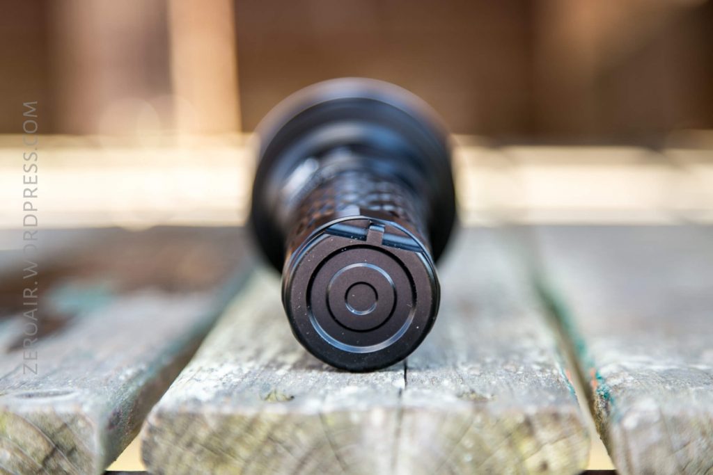 A close-up view of the tail end of a black flashlight resting on a weathered wooden surface, with the background blurred.