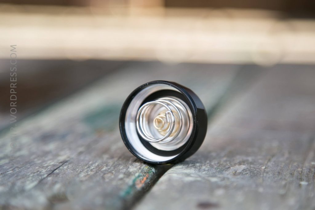 A close-up of a metal spring inside a black circular casing is placed upright on a weathered wooden surface. The background is blurred.
