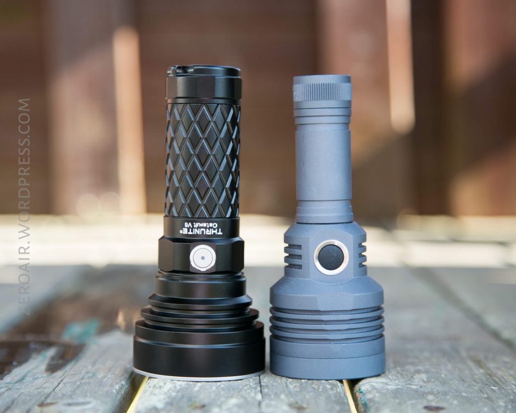 Two flashlights standing upright on a wooden surface outdoors; the left flashlight is shorter, black, and has a textured grip, while the right flashlight is taller, grey, and has a smooth finish.