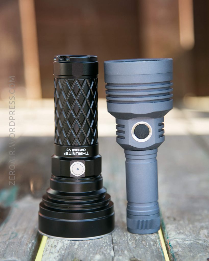 Two flashlights stand upright on a wooden surface. The flashlight on the left is black with a textured grip, while the one on the right is gray with a smooth body and a larger head.