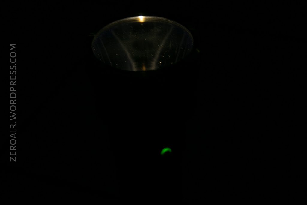A flashlight with a faintly illuminated lens and a small green light on its body is partially visible in a dark setting. The surrounding background is mostly black. The text “ZEROAIR.WORDPRESS.COM” is written vertically on the left.