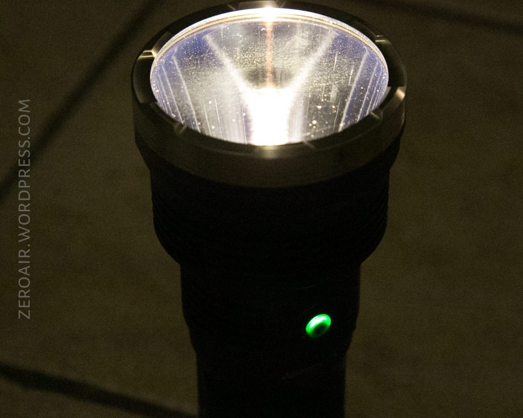 A lit flashlight with a metal bezel and a green power button stands on a tiled floor, emitting a bright light upwards. The text ZEROAIR.WORDPRESS.COM is visible along the left edge.