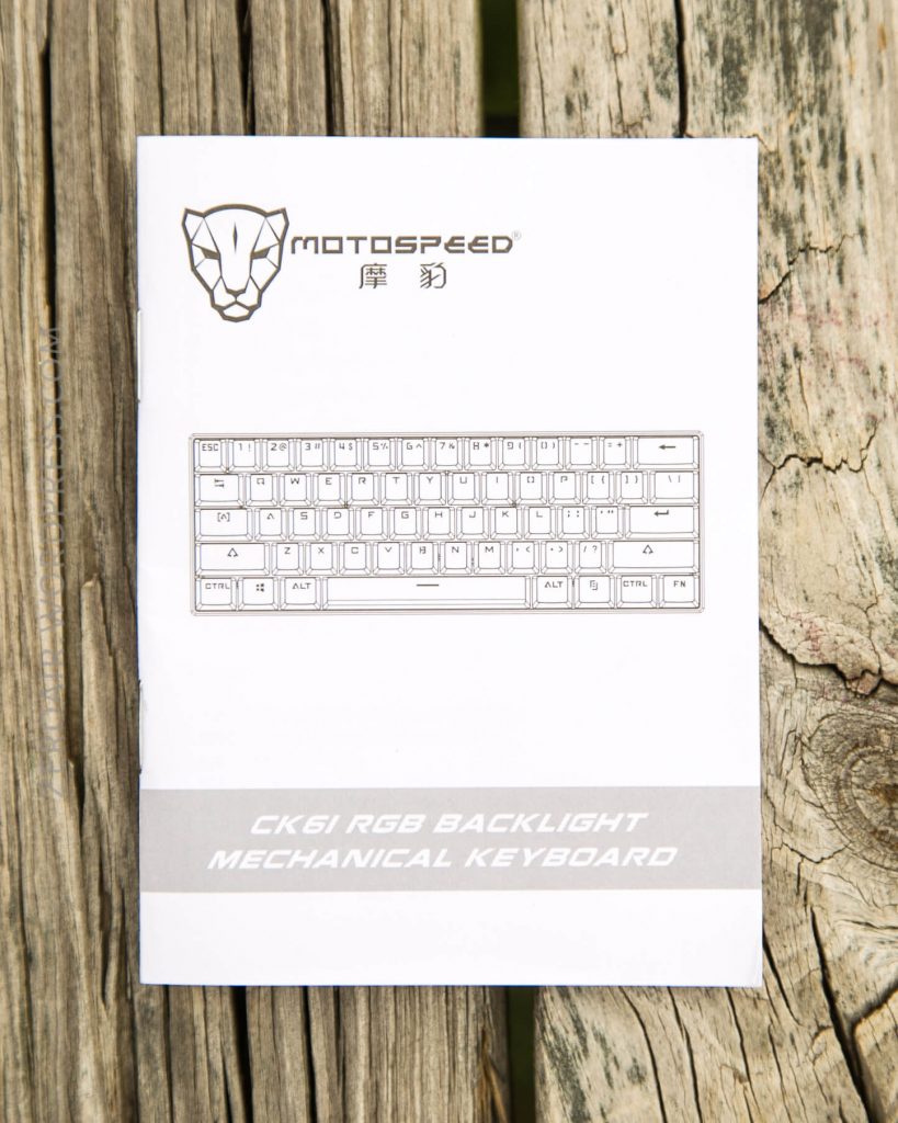 A white user manual for the Motospeed CK61 RGB Backlight Mechanical Keyboard rests on a weathered wooden surface. The manual features a keyboard diagram and product branding on the cover.
