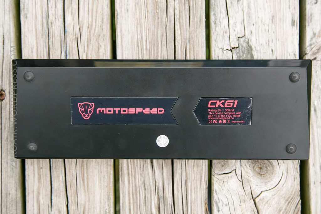 The underside of a black Motospeed CK61 keyboard is shown on a wooden surface. Visible are the Motospeed logo, product label, rubber feet, and screws.