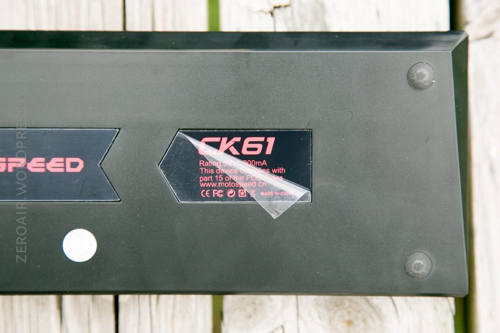 A close-up of the back of a black mechanical keyboard, showing a label with the model GK61 and product information partially covered by a clear piece of tape. The keyboard rests on a wooden surface.