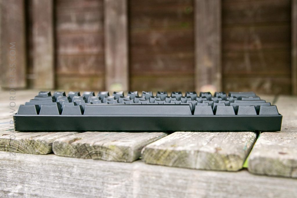 A black computer keyboard is placed on a weathered wooden bench outdoors, with a blurred wooden wall in the background. The photo is taken from a low, side angle.