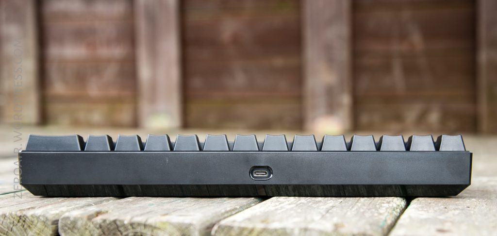 A close-up side view of a black mechanical keyboard showing the keycaps and a USB-C port, placed on a weathered wooden surface with a blurred wooden background.