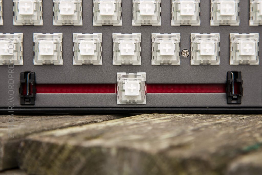 Close-up of a mechanical keyboard without keycaps, showing transparent switches and a visible red stabilizer bar underneath the spacebar position, placed on a wooden surface.