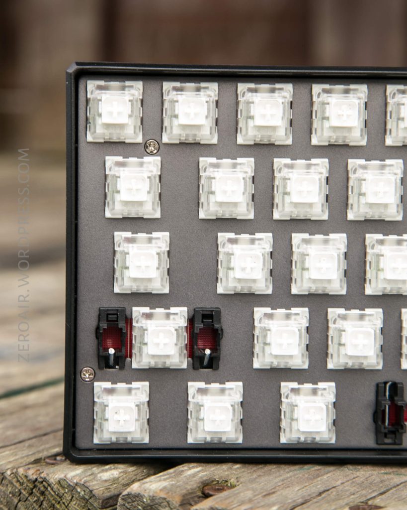 A close-up of a mechanical keyboard without keycaps, showing several white switches and three red switches in the lower left area, mounted in a black frame, placed on a wooden surface.