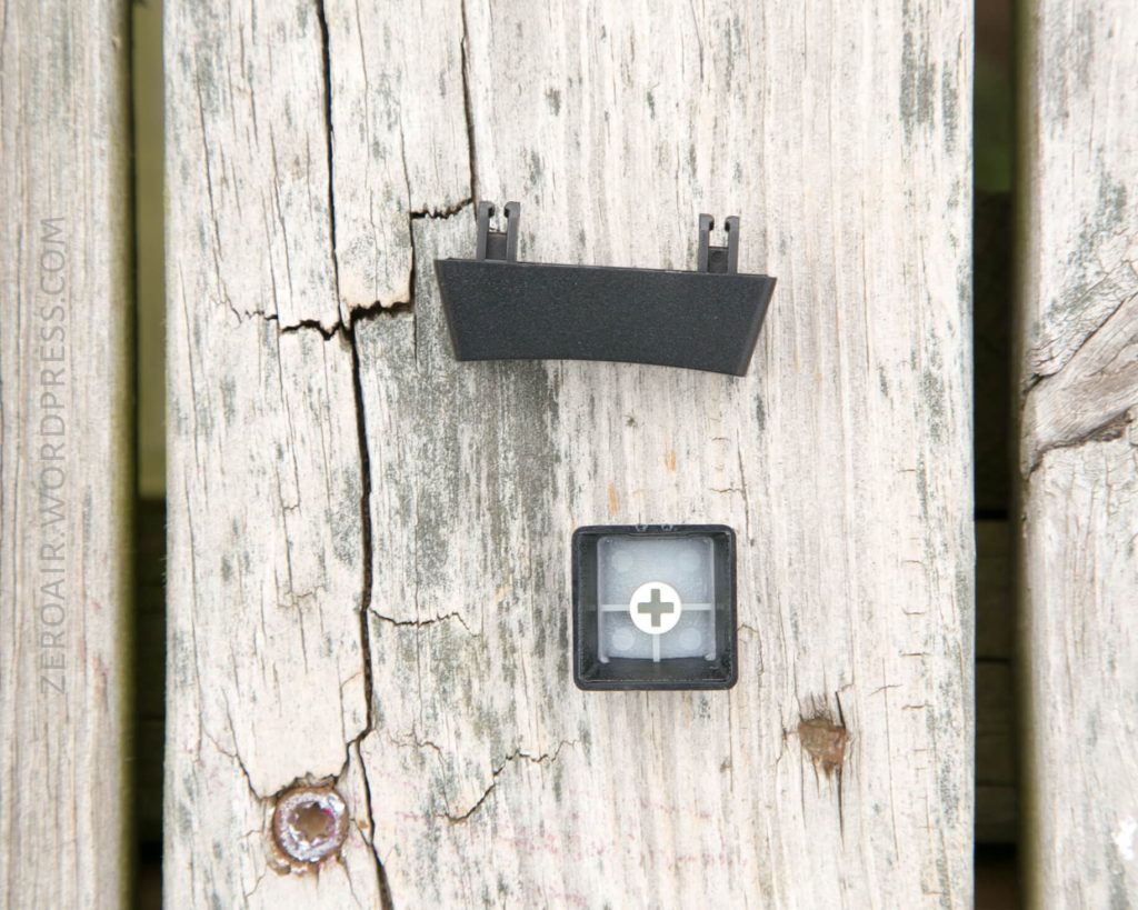 A black keyboard keycap and a keycap stabilizer are placed on a weathered wooden surface. The keycap shows the underside with a cross-shaped connector visible.