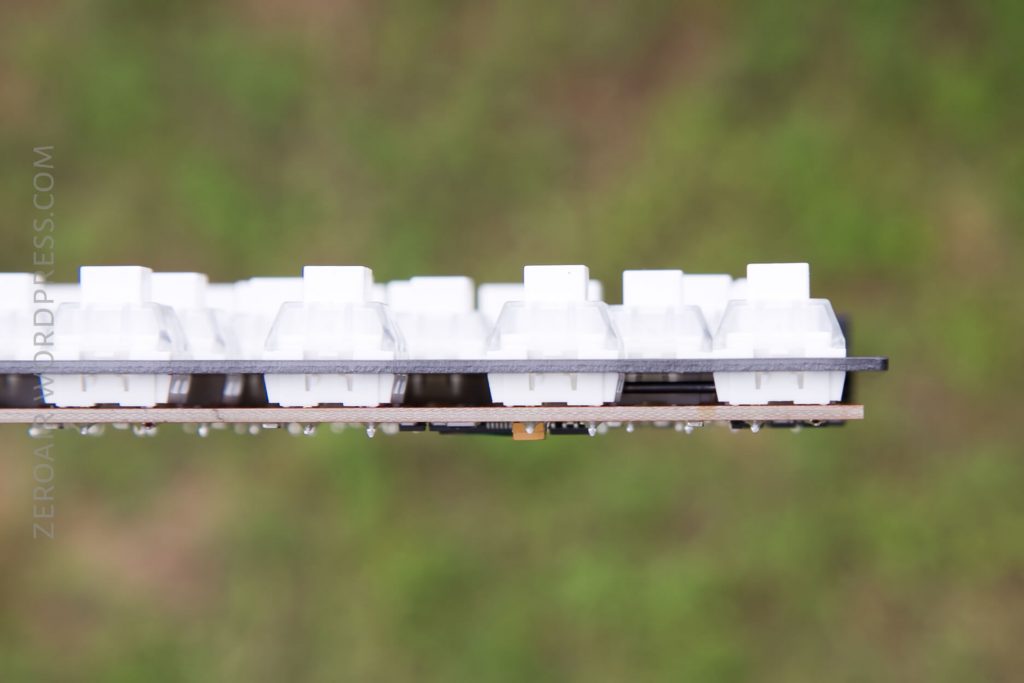 Side view of a keyboard PCB with white mechanical key switches installed, against a blurred green background. The switches and circuit board are visible, with no keycaps attached.