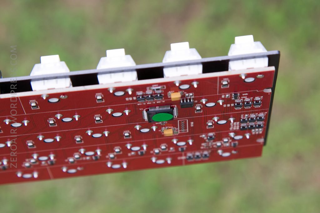 A close-up of a red printed circuit board with white key switches and various electronic components, photographed outdoors with a blurred green background.
