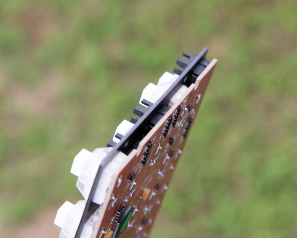 A close-up view of the edge of a brown circuit board with white and black plastic components, against an out-of-focus grassy background.