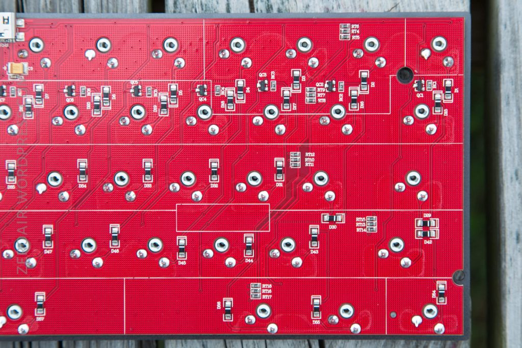 A close-up of a red printed circuit board with multiple solder points, mounted components, and traces, photographed on a wooden surface. No electronic components are installed in the visible slots.