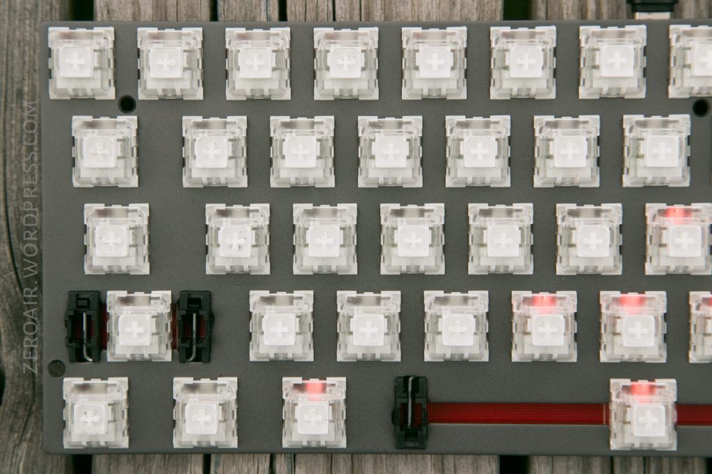 A close-up view of a mechanical keyboard plate with multiple clear key switches installed, some featuring red stems, on a wooden surface. The switches are unkeyed and the board is partially assembled.