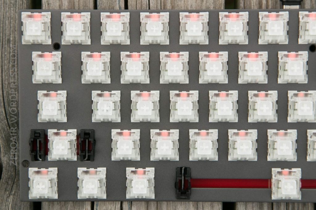 A close-up view of a mechanical keyboard plate with several translucent switch housings installed, some with red stems and a few with black stems, set on a wooden surface.