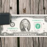A U.S. two-dollar bill is placed on a wooden surface. A black rectangular object partially obscures the left side of the bill. The portrait of Thomas Jefferson is visible on the bill.