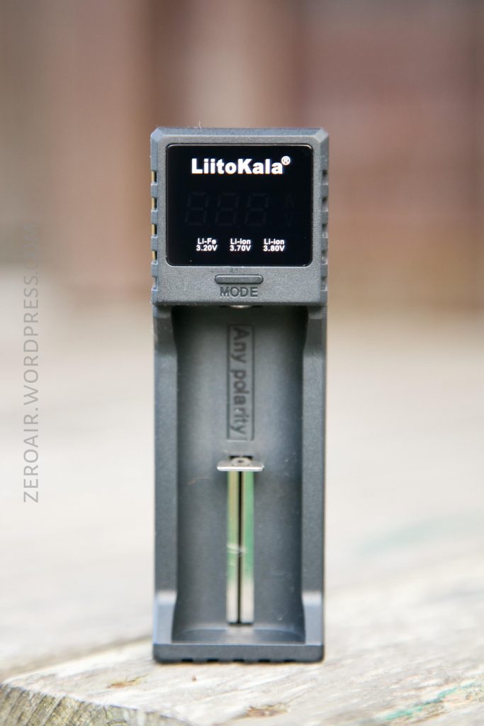 A black LiitoKala battery charger is standing upright on a wooden surface. The charger display is off, and a single charging slot is visible below the screen. The MODE button is located below the display.