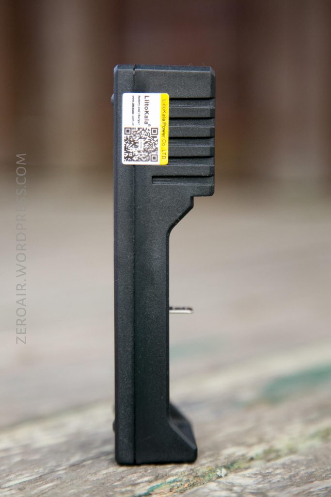 A black electronic device, standing upright on a wooden surface, with a yellow and white label and a QR code on its side. A metal pin protrudes from the center of the device.