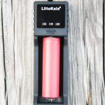 A LiitoKala battery charger is holding a single red cylindrical battery. The device is placed on a weathered wooden surface. The chargers display shows battery types and a MODE button is visible below the battery.