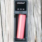 A red cylindrical battery is placed in a black LiitoKala charger on a wooden surface. The chargers display shows different voltage options, and the word MODE is printed below the battery compartment.