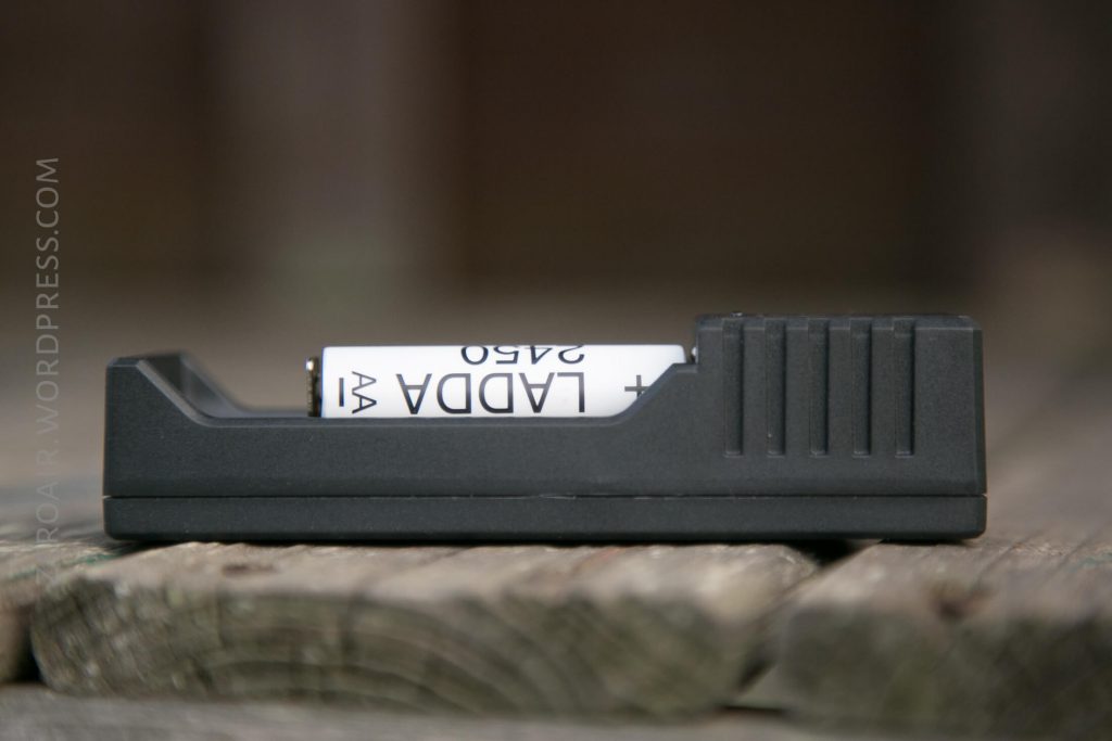 A close-up, side view of a black battery charger holding a white AA rechargeable battery labeled “LADDA 2450,” placed on a wooden surface.