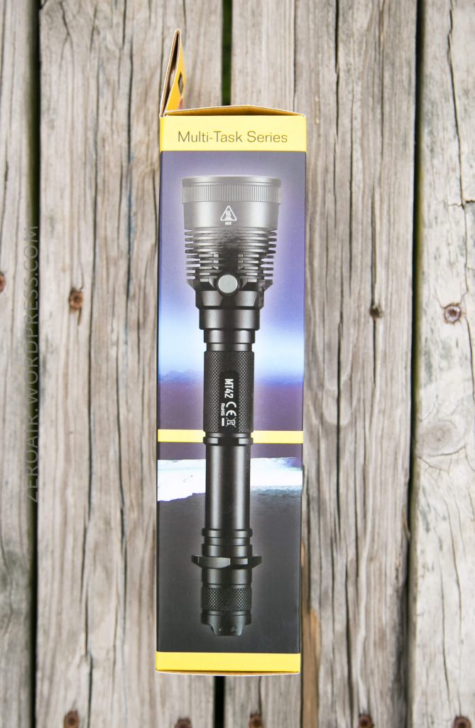 A yellow and black flashlight box labeled Multi-Task Series stands upright on a weathered wooden surface. The box displays an image of a large black flashlight on the front.