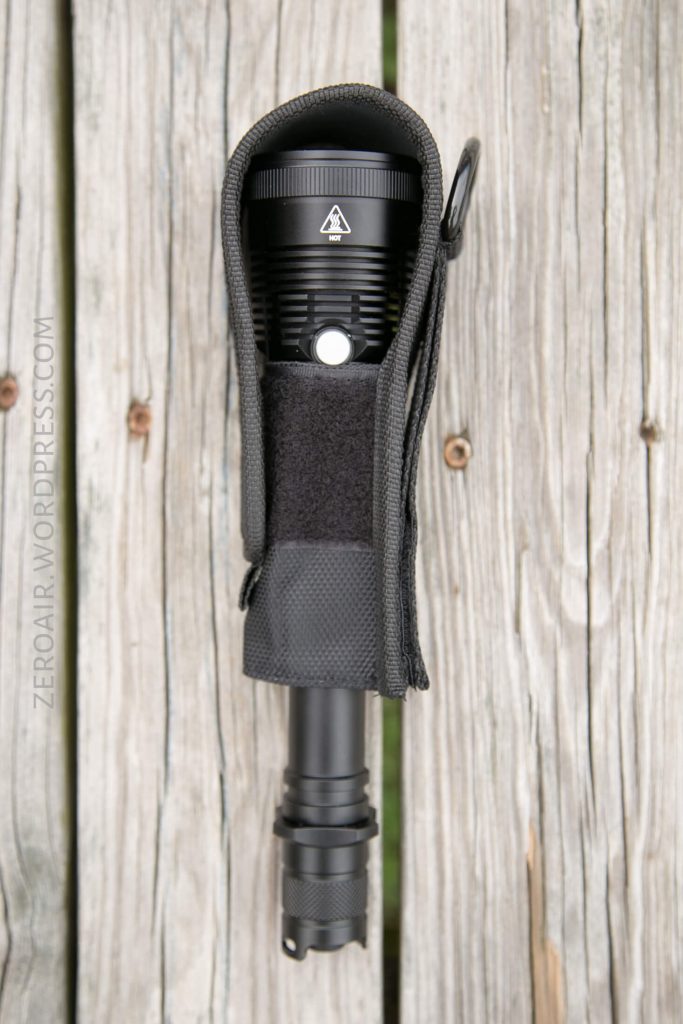 A black flashlight partially enclosed in a black fabric holster is placed vertically on a wooden surface. The holster flap is open, and the text ZEROAIR.WORDPRESS.COM is visible along the left side.