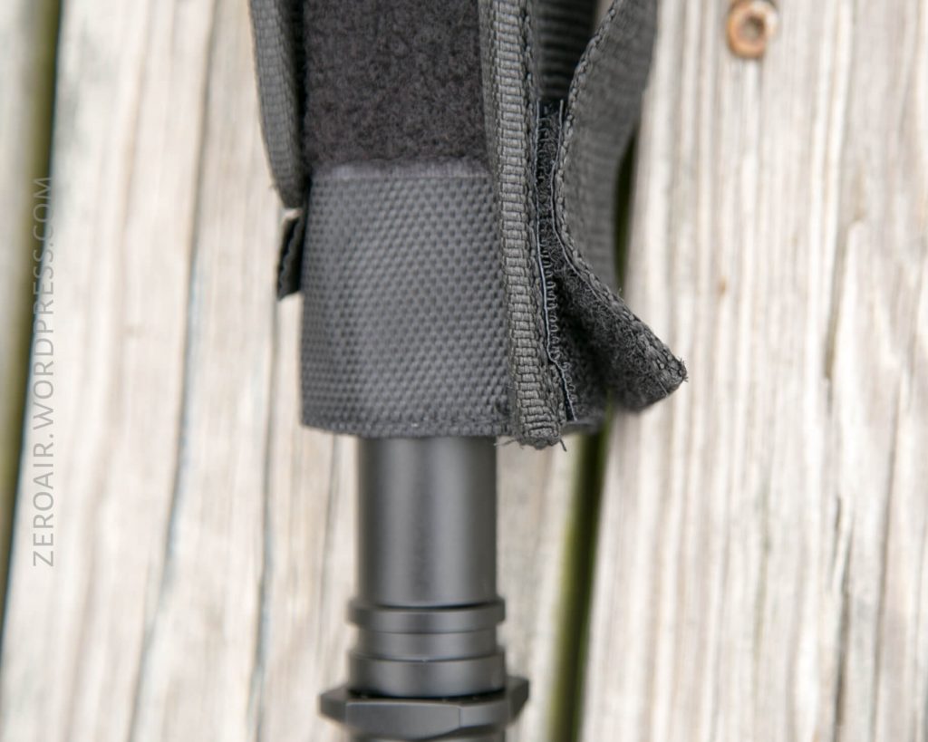 A close-up of a black nylon strap and fabric partially covering a metal cylindrical object, placed on a weathered wooden surface. The stitching and texture of the materials are visible.