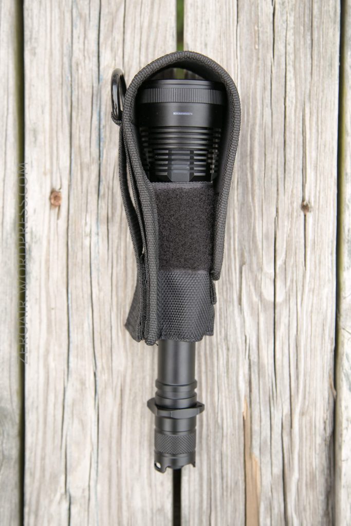 A black flashlight in a fabric holster with a Velcro closure is attached to a weathered wooden surface. The holster is open, revealing the flashlight’s lens and bezel.