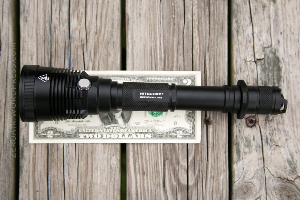 A black Nitecore flashlight is placed on top of a two-dollar bill on a wooden surface, showing the size comparison between the flashlight and the bill.