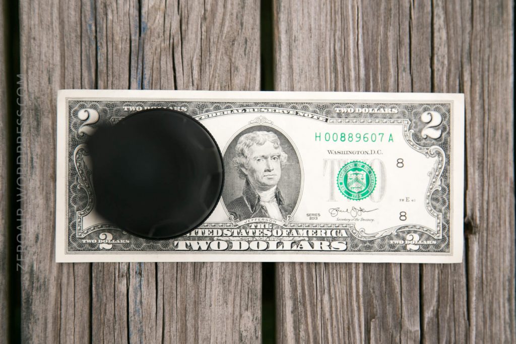 A two-dollar bill is placed on a wooden surface with a black drink coaster covering the left side of the bill.