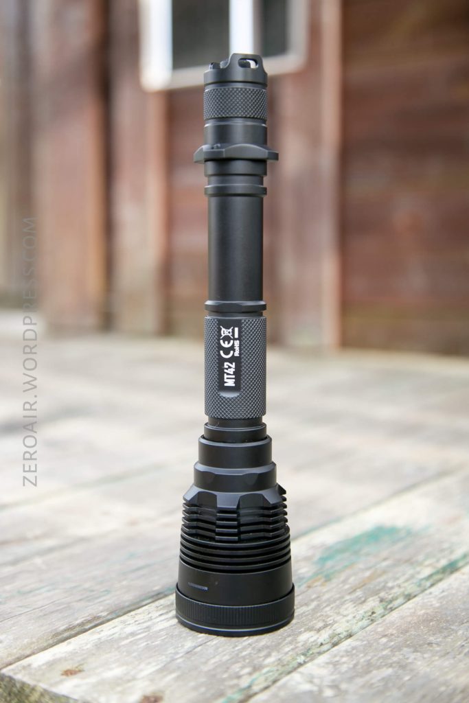 A black, heavy-duty flashlight stands upright on a weathered wooden surface with a blurred brown and white background. The flashlight is labeled MT25 X7 and features a textured grip and ridged head.